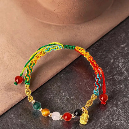 A multicolor braided cord bracelet with gold-toned links, colorful round beads, and a small gold charm, displayed on a dark textured surface with a beige stone prop.