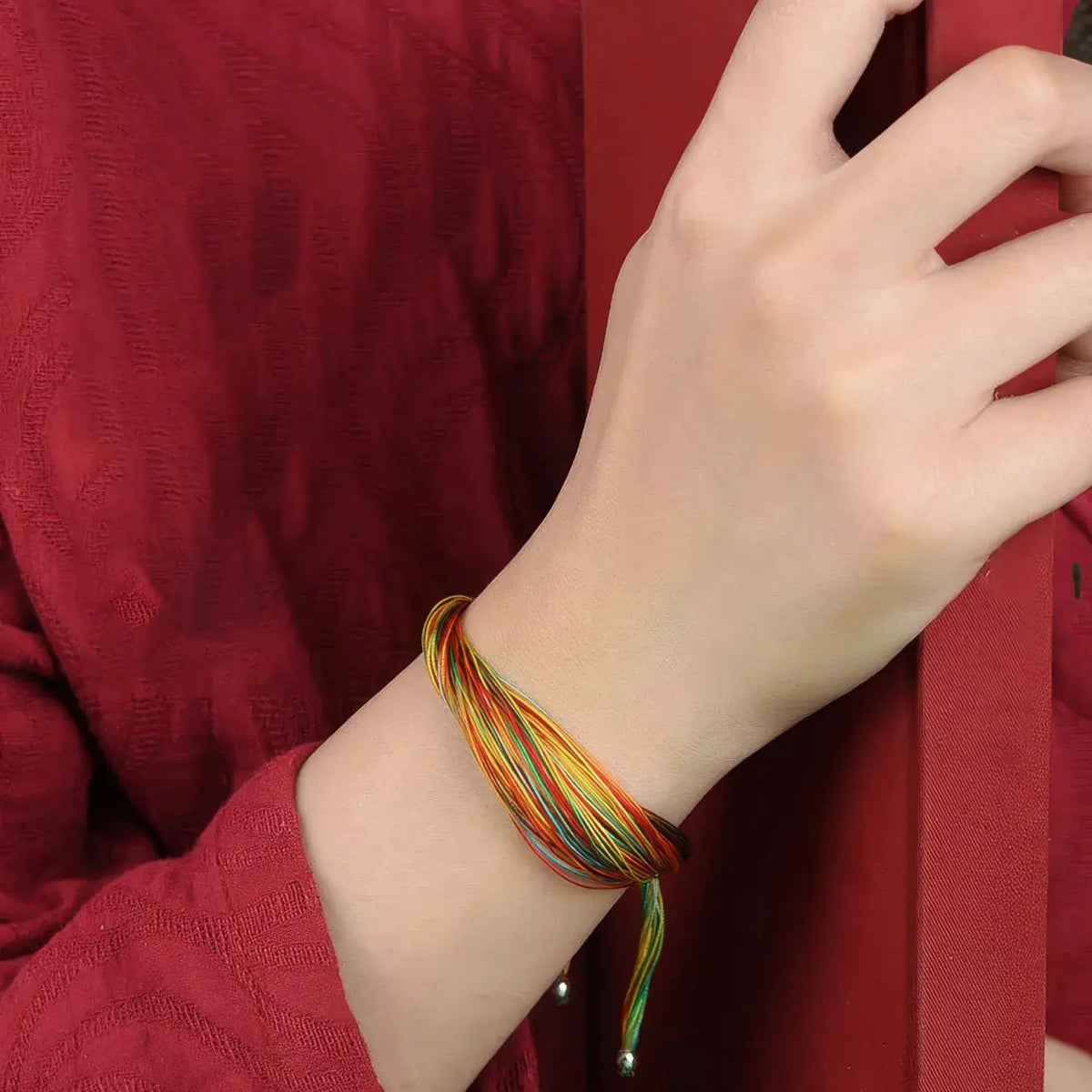 A close-up of a person's wrist wearing a multi-colored braided cord bracelet with small metallic beads, set against a red textured garment.