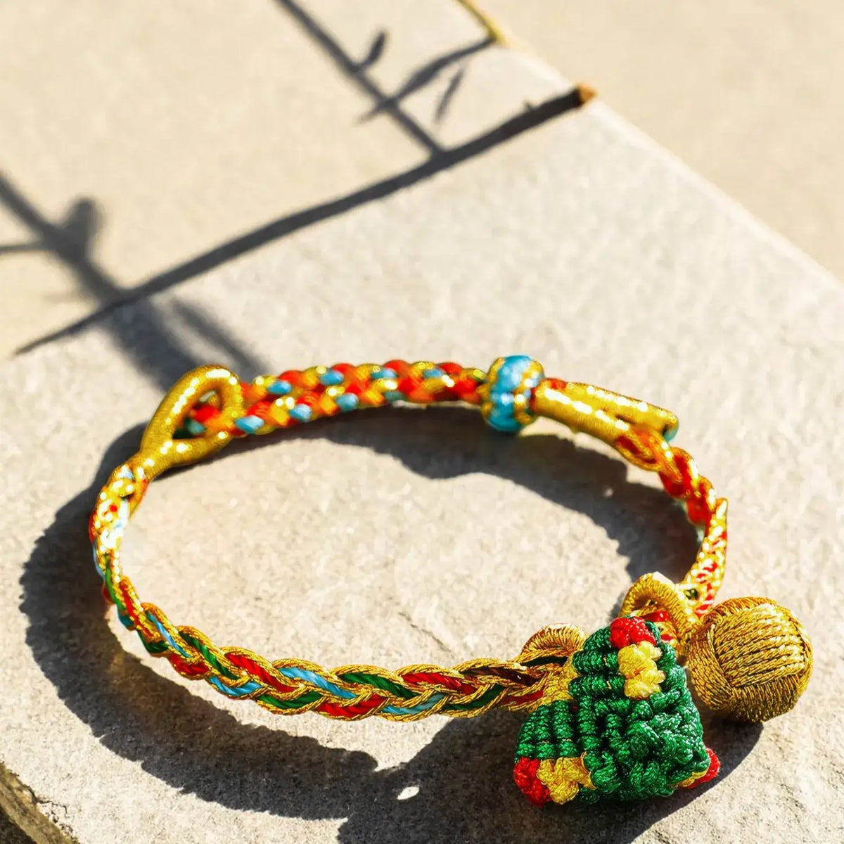 A handmade braided bracelet with gold-tone metal accents, a green and red knotted ornament, and a gold bell, photographed on a stone surface in bright sunlight.