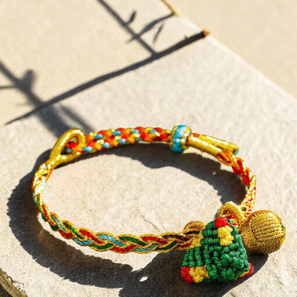 A handmade braided bracelet with gold-tone metal accents, a green and red knotted ornament, and a gold bell, photographed on a stone surface in bright sunlight.
