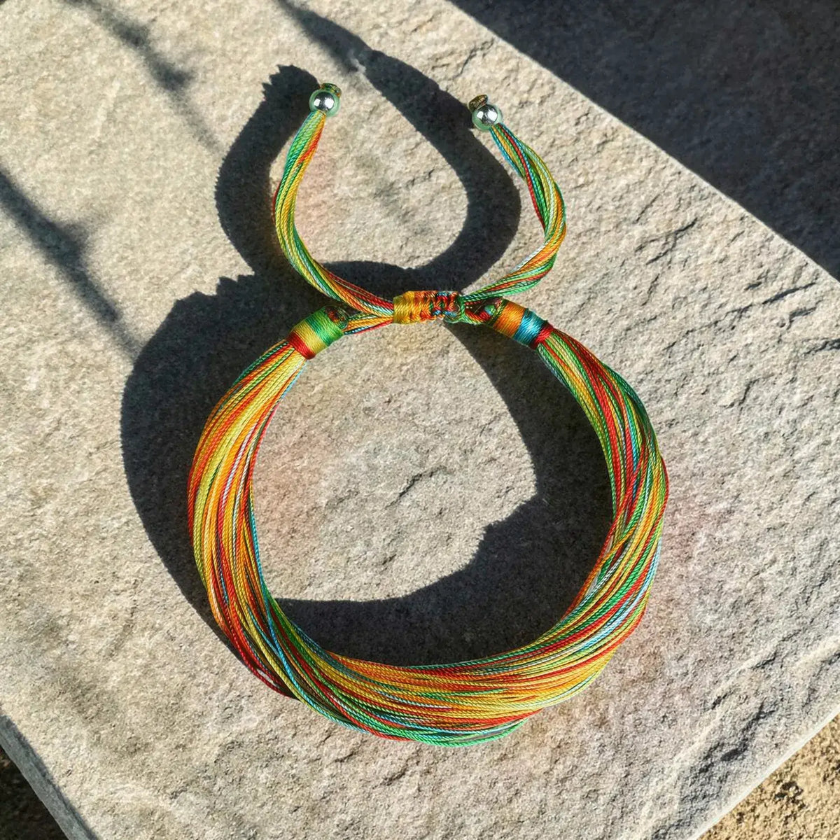 A multicolor braided cord bracelet with multiple strands of red, orange, yellow, green, and blue cords, laid out on a textured gray stone surface in direct sunlight.