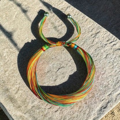 A multicolor braided cord bracelet with multiple strands of red, orange, yellow, green, and blue cords, laid out on a textured gray stone surface in direct sunlight.