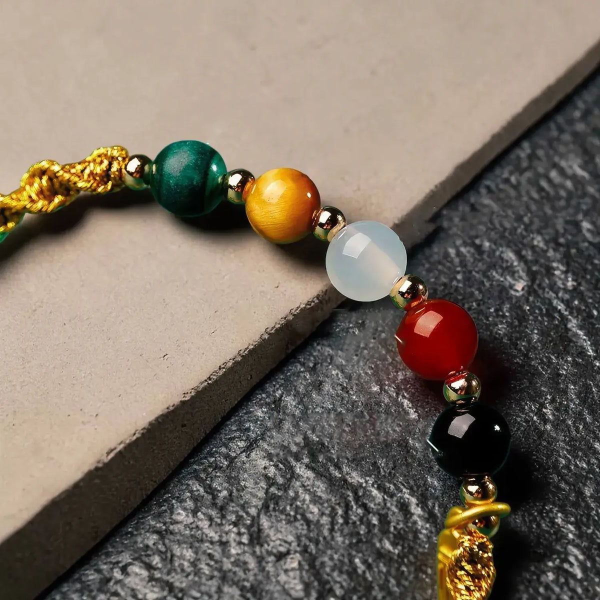 A close-up of a beaded bracelet featuring polished gemstone-like beads in green, orange, white, red, black, and gold, with a gold cord on a neutral stone background.