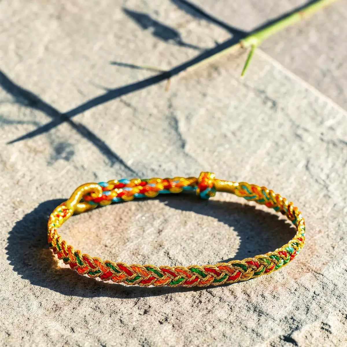 A braided Chinese friendship bracelet with gold, red, green, and blue threads, resting on a textured gray stone surface under bright sunlight.
