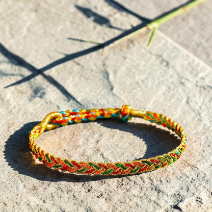 A braided Chinese friendship bracelet with gold, red, green, and blue threads, resting on a textured gray stone surface under bright sunlight.