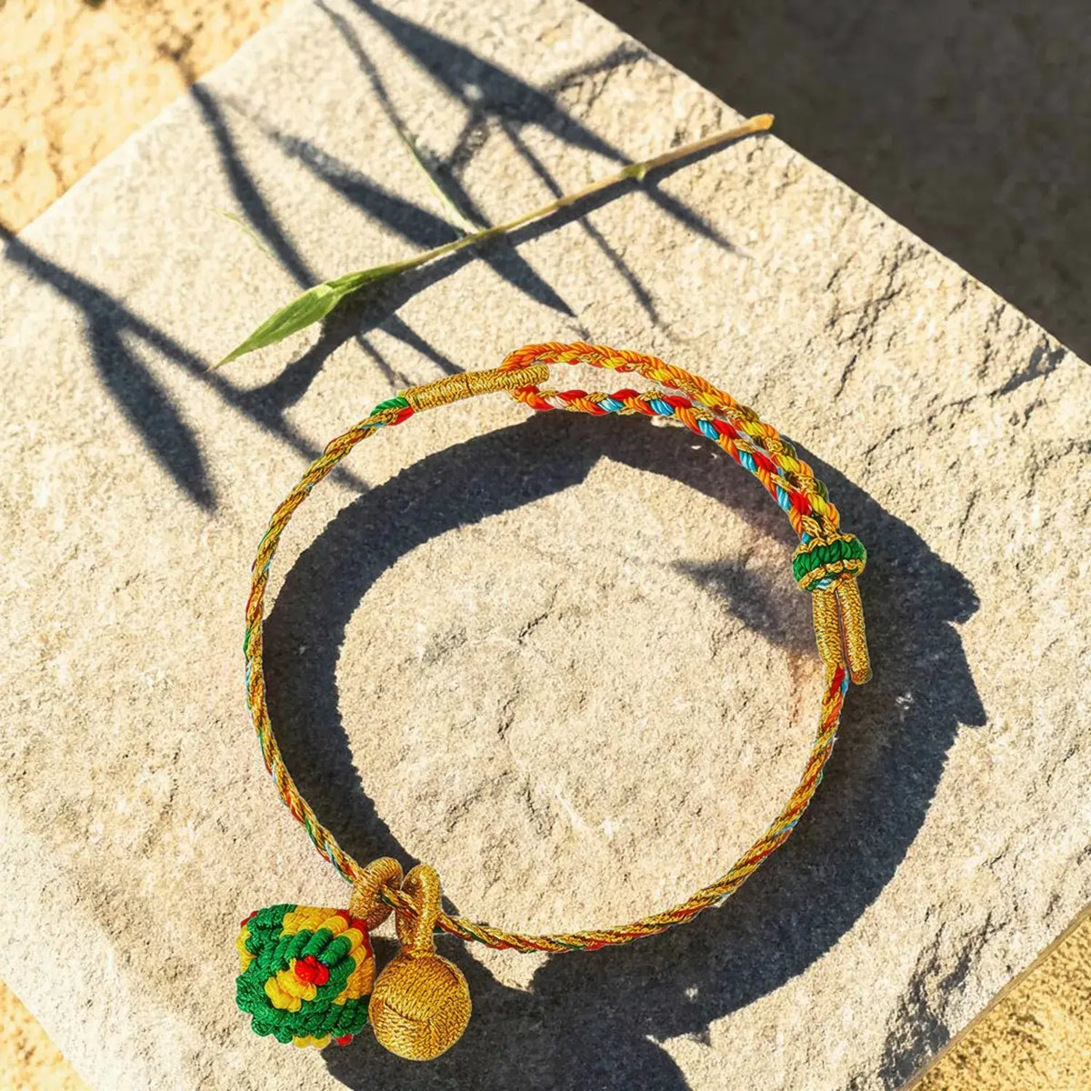 A braided cord bracelet with a small hanging ornament, photographed on a stone surface in bright sunlight. The bracelet is multicolored, with gold-toned braided sections, green and blue woven details, and a green-and-yellow decorative charm.