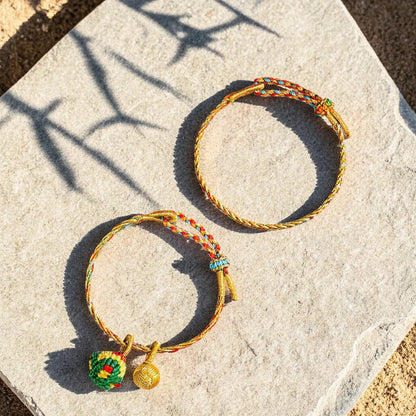 Two braided cord bracelets with small hanging charms, displayed on a light stone surface in warm sunlight with leaf shadows.