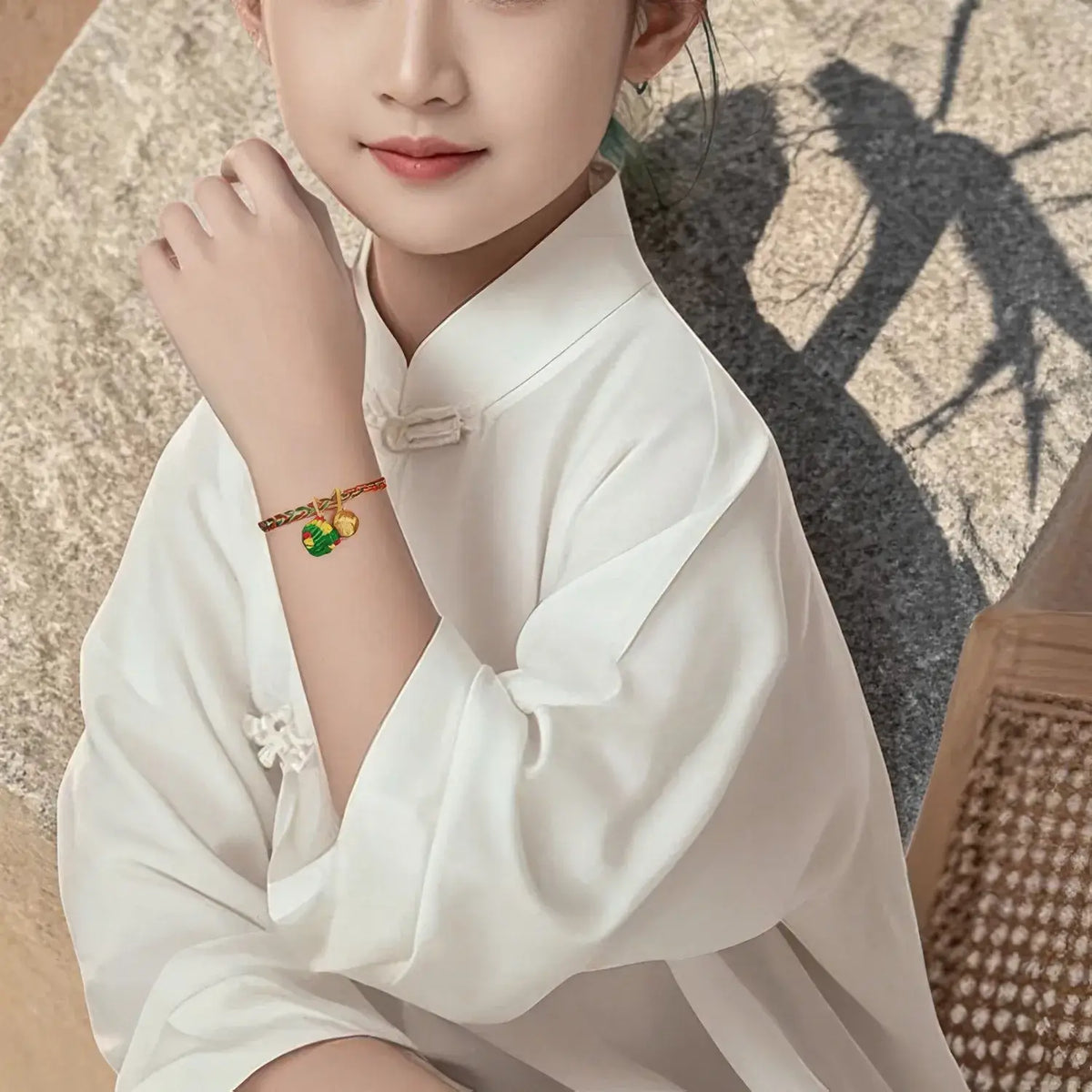 A child wearing a white traditional-style top and a gold bracelet with green and red decorative charms, posed outdoors against a textured stone wall with a strong shadow.