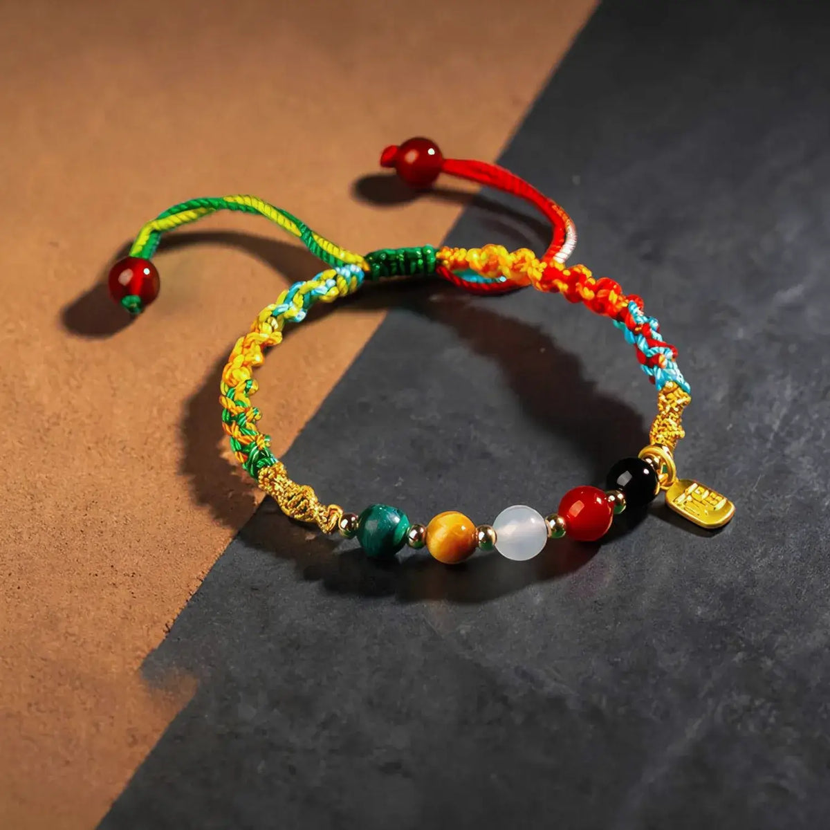 Two colorful Chinese-style cord bracelets with gold-plated hardware and gemstone beads, displayed on a split brown and dark gray background.