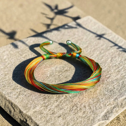 A multicolor adjustable cord bracelet with silver metal end caps, resting on a textured stone slab outdoors under direct sunlight.