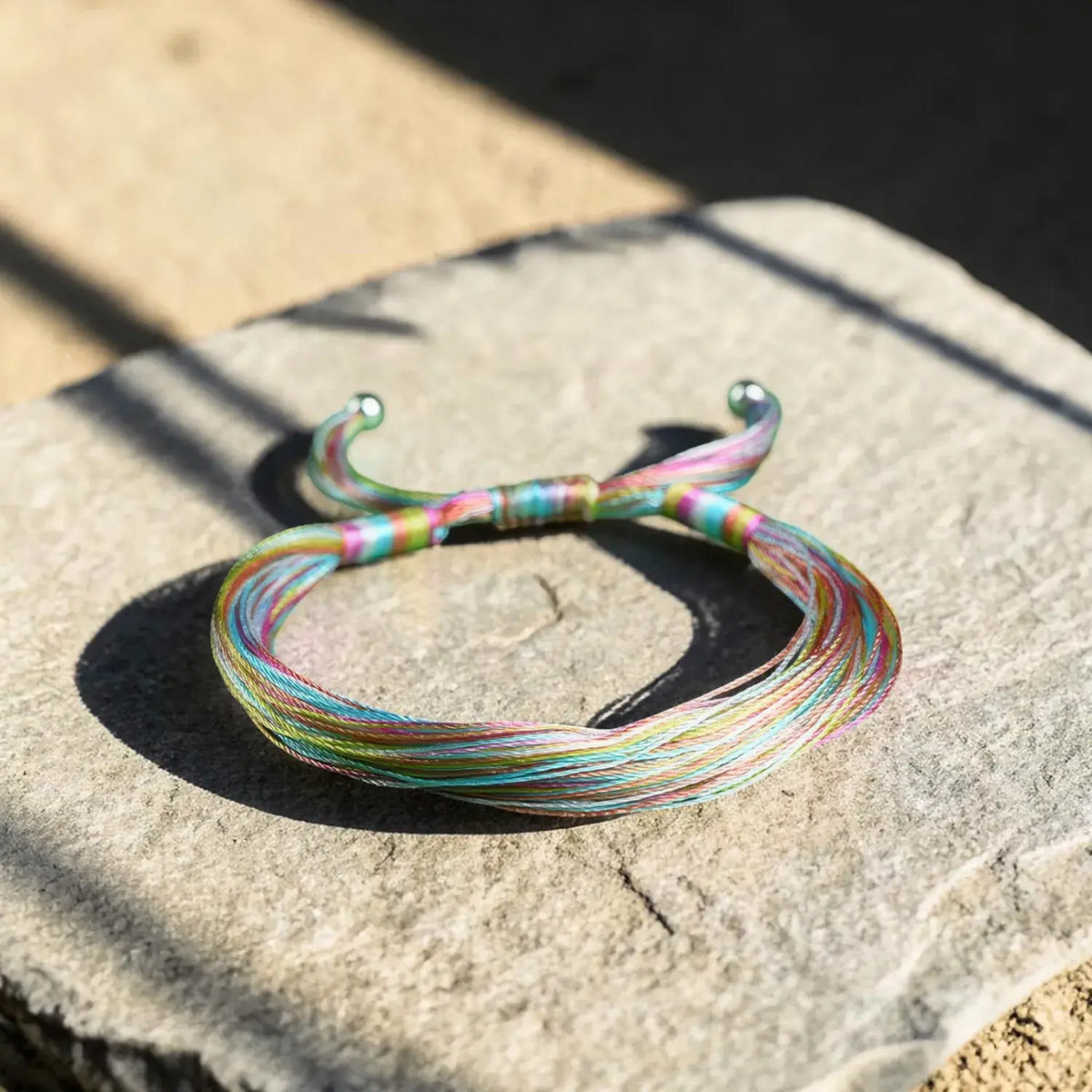 A thin multicolor braided bracelet with silver-toned metal ends, resting on a textured stone surface in natural sunlight.