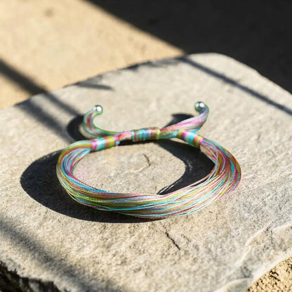 A thin multicolor braided bracelet with silver-toned metal ends, resting on a textured stone surface in natural sunlight.