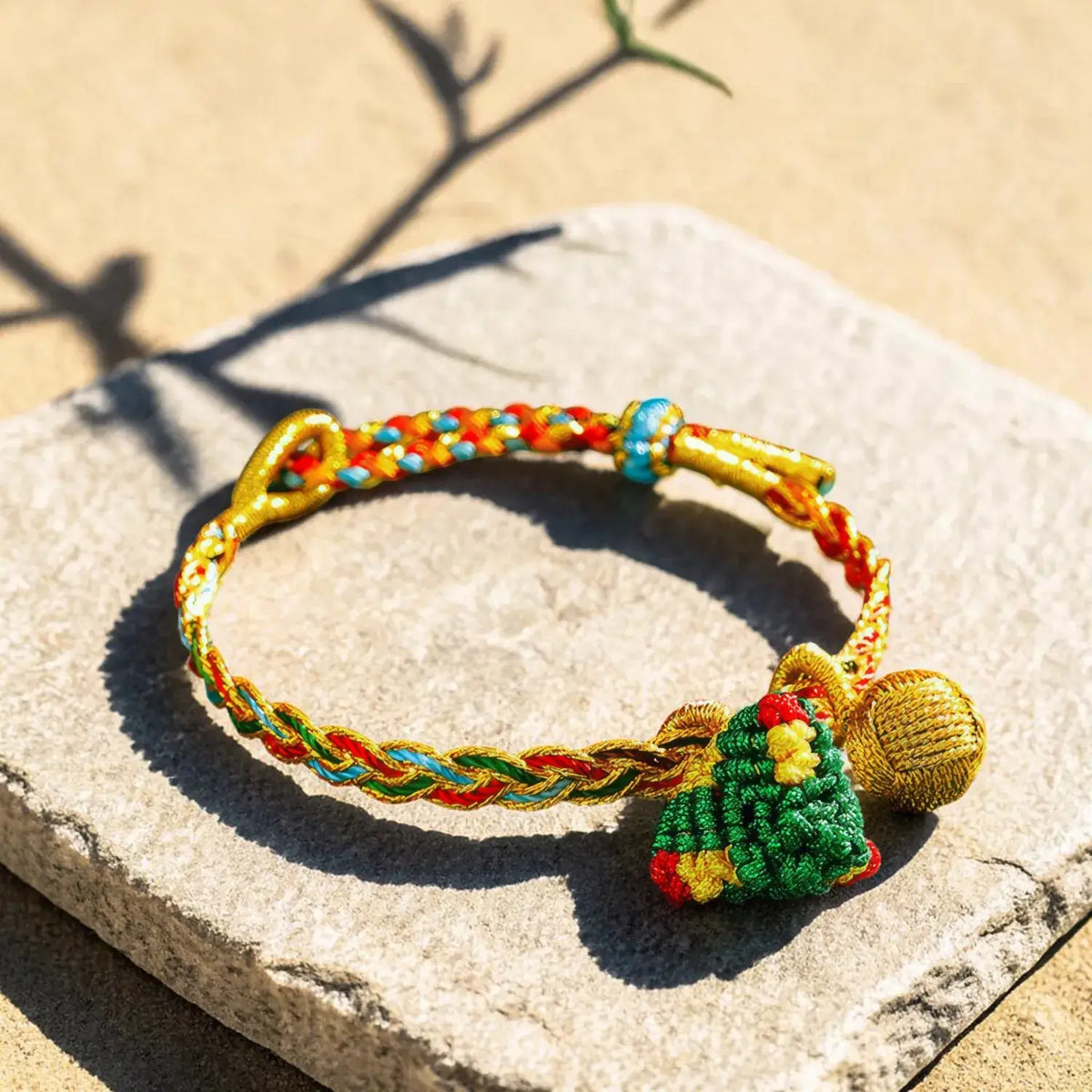 A braided and knotted bracelet with a small green Christmas tree charm and a gold bell, displayed on a stone slab in bright sunlight.