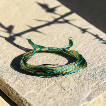 A handmade braided bracelet made of thin green, teal, and gold threads, resting on a textured stone surface under bright sunlight with sharp shadows.