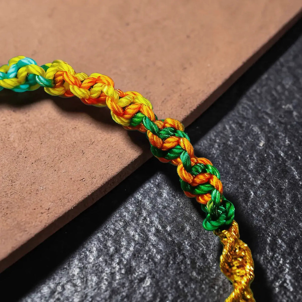 A close-up of a braided cord bracelet or necklace with woven segments in teal, yellow, orange, and green, laid diagonally across a brown tile and dark textured surface.