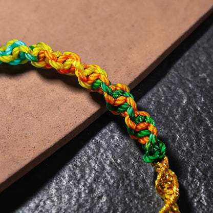 A close-up of a braided cord bracelet or necklace with woven segments in teal, yellow, orange, and green, laid diagonally across a brown tile and dark textured surface.
