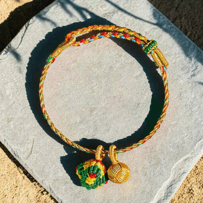 A braided gold-toned cord bracelet with colorful red, blue, and green accents, featuring two hanging charms including a small woven green and yellow ornament and a gold bead, displayed on a light gray stone slab outdoors.