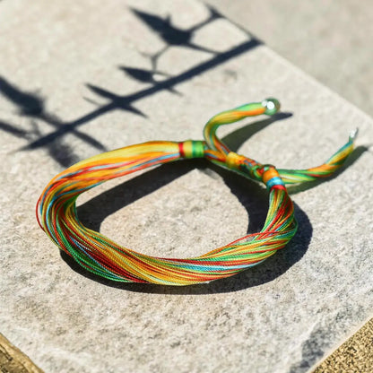 A multicolor braided cord bracelet with yellow, green, orange, and red strands, resting on a textured stone surface under bright sunlight with leaf shadows.