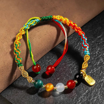 A multicolor braided cord bracelet with red, yellow, green, blue, and orange cords, decorated with colorful round beads and a gold-tone charm, displayed on a dark textured surface.