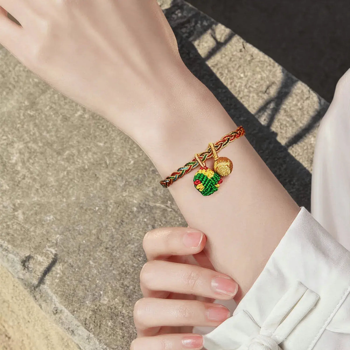 A close-up of a person's wrist wearing a braided red and green cord bracelet with two small charms: one gold-toned leaf and one green and red patterned heart charm.