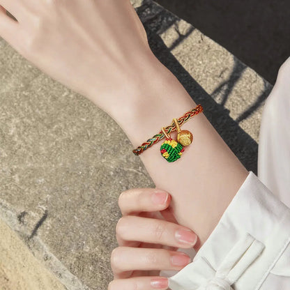 A close-up of a person's wrist wearing a braided red and green cord bracelet with two small charms: one gold-toned leaf and one green and red patterned heart charm.