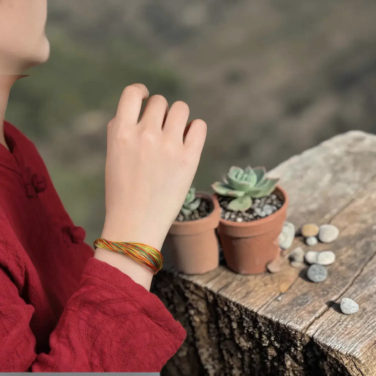 A person wearing a red long-sleeve top and a thin braided bracelet with orange, yellow, and green threads, sitting beside two small potted succulents on a rustic wooden surface.
