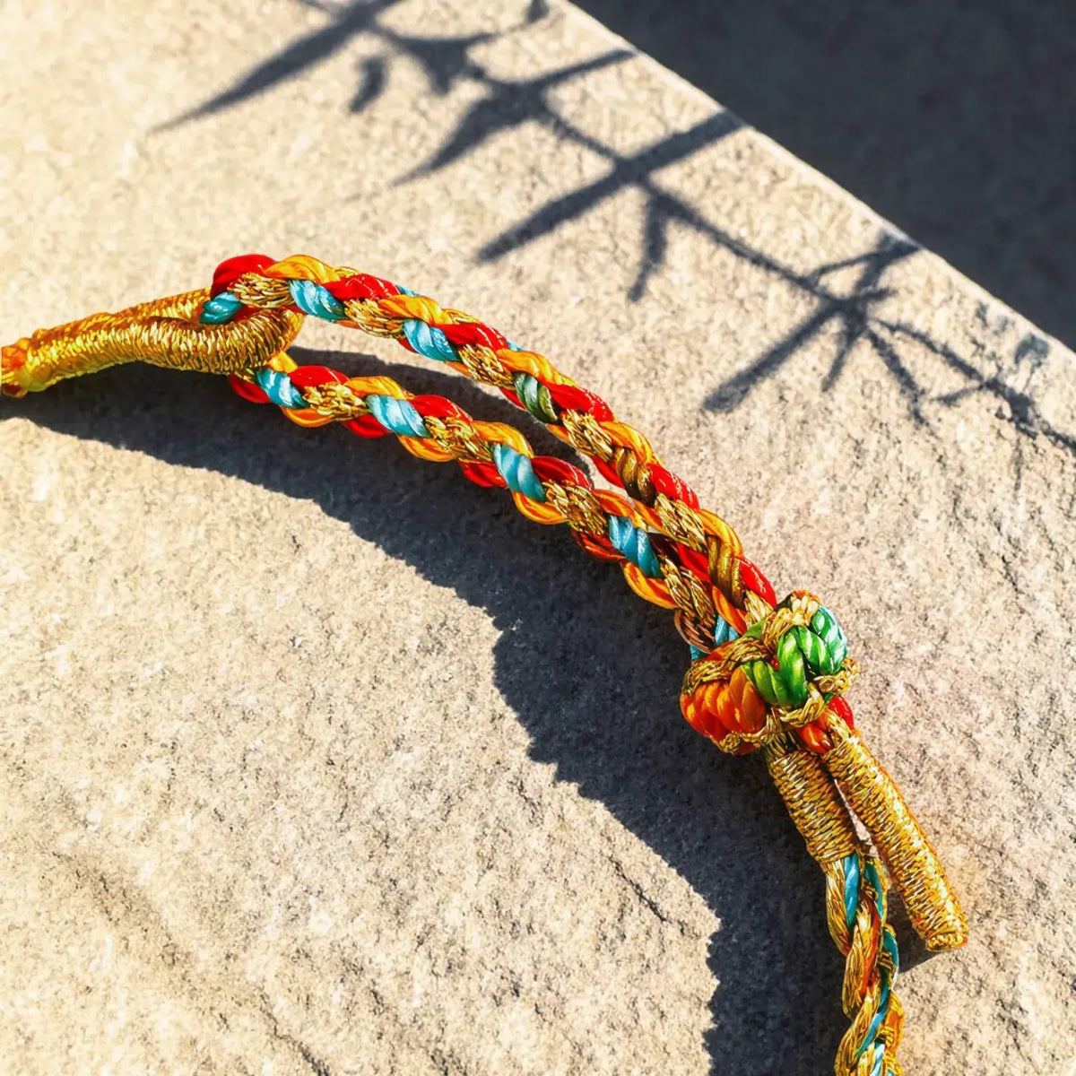 A close-up of a braided cord bracelet or tassel accessory made of gold, red, orange, blue, and green threads, resting on a textured stone surface with sharp shadows.