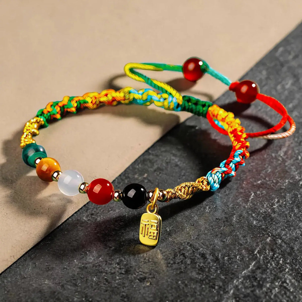 A close-up of a braided cord bracelet with colorful woven cords, round beads in red, green, orange, white, and black, and a small gold charm engraved with Chinese characters, resting on a split beige and dark gray surface.