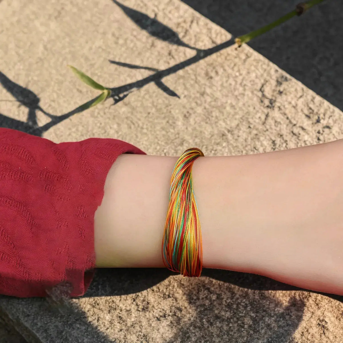 A close-up of a person's wrist wearing a multi-colored braided cord bracelet, with a red sleeve visible and a stone surface in the background.