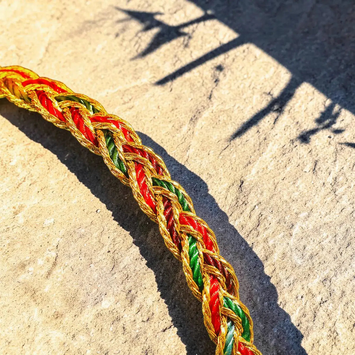 A close-up of a braided rope or cord in red, green, and gold, laid diagonally across a light beige stone surface with strong sunlight and shadows.