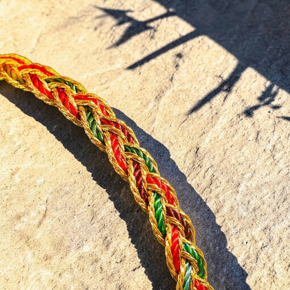 A close-up of a braided rope or cord in red, green, and gold, laid diagonally across a light beige stone surface with strong sunlight and shadows.