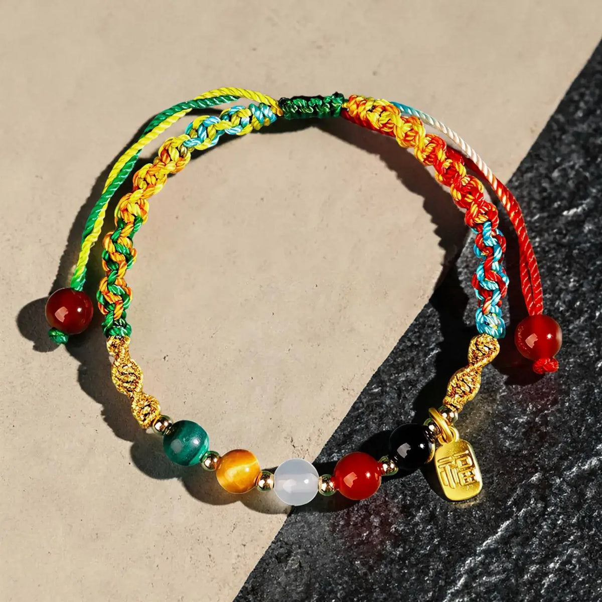 A handmade multi-colored braided cord bracelet with colorful beads and a gold charm, displayed on a split beige and dark gray surface.