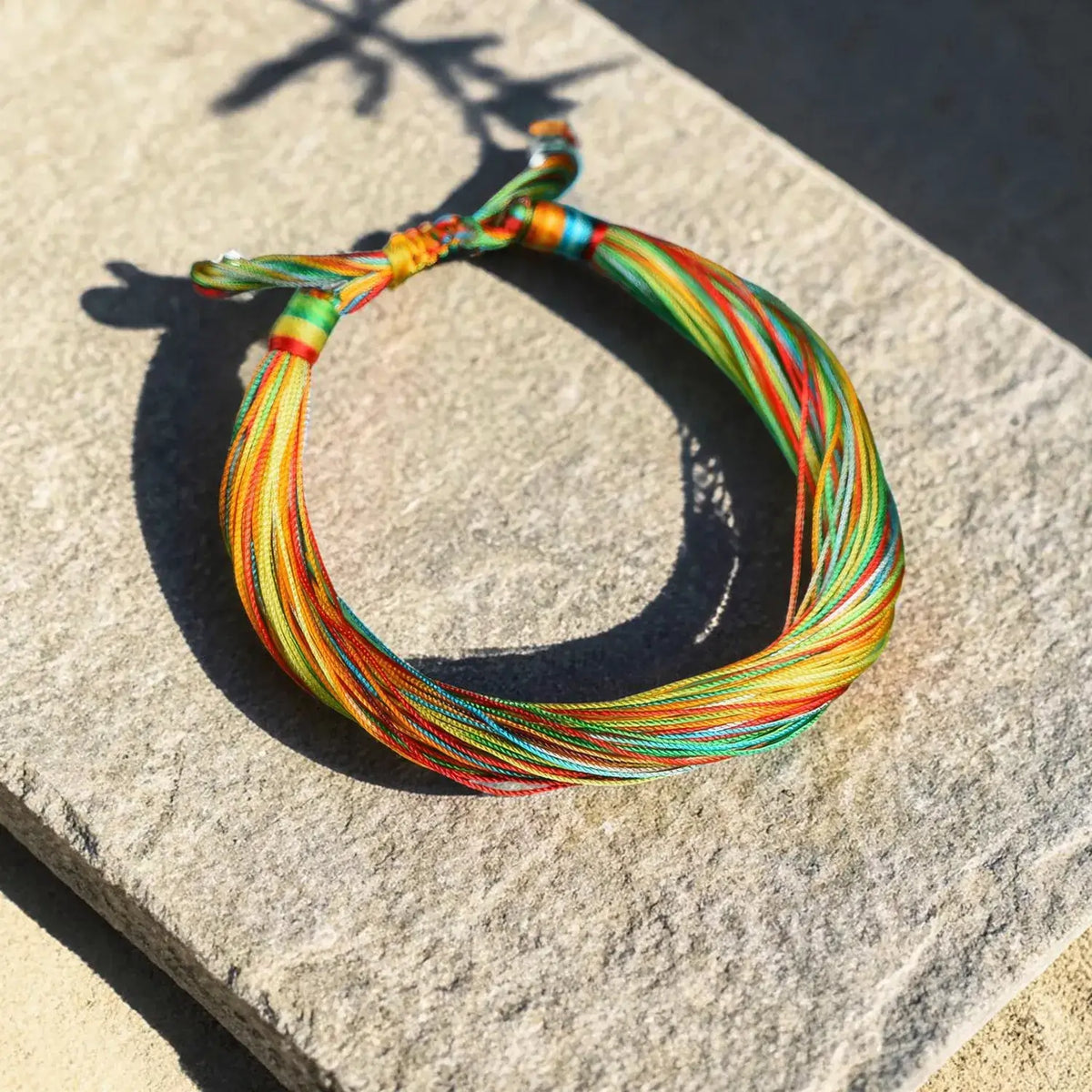 A multicolor braided cord bracelet with red, yellow, green, and orange threads, tied in a knot and displayed on a textured gray stone surface in direct sunlight.