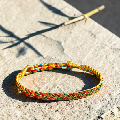 A braided cord bracelet with red, yellow, green, and blue threads, displayed on a textured stone surface in bright sunlight.