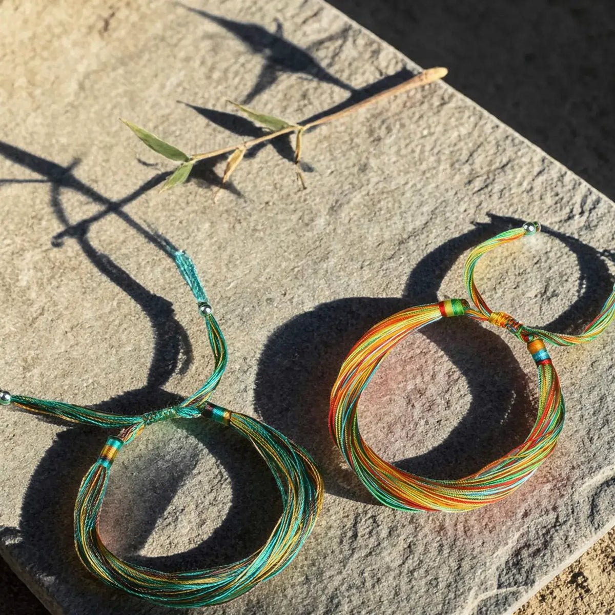 Two handmade multi-strand cord bracelets with colorful threads and small bead accents, displayed on a textured stone surface in direct sunlight.