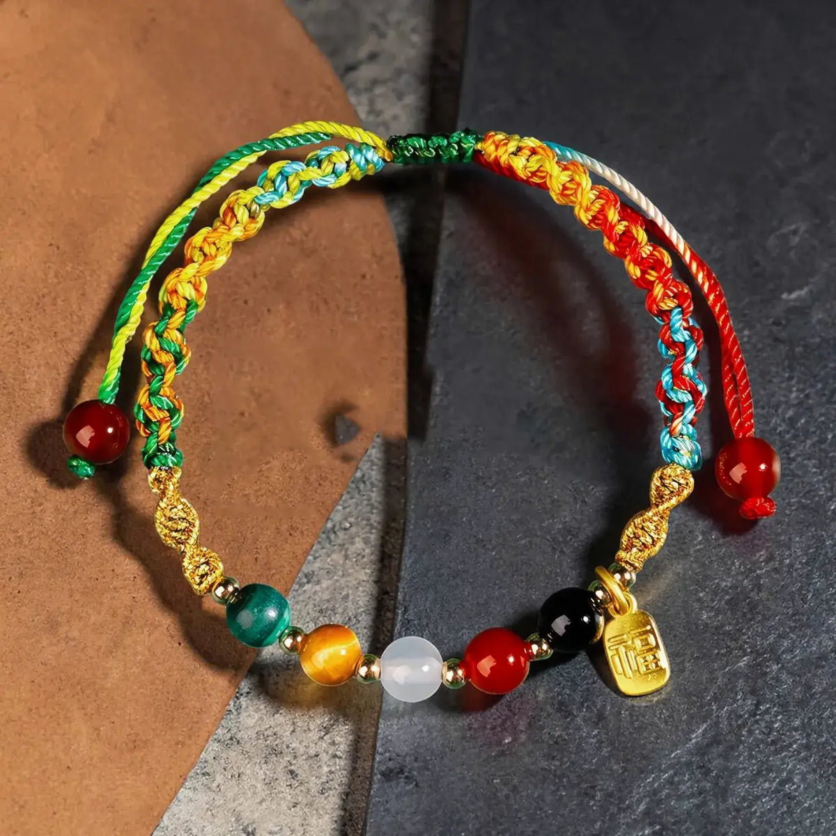 A handmade multicolor braided cord bracelet with red, green, yellow, blue, and orange cords, decorated with colorful beads and a small gold charm, displayed on a split brown and dark gray background.