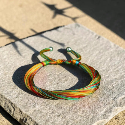 A multicolor braided cord bracelet with green metal toggle sliders, resting on a textured gray stone slab in direct sunlight.