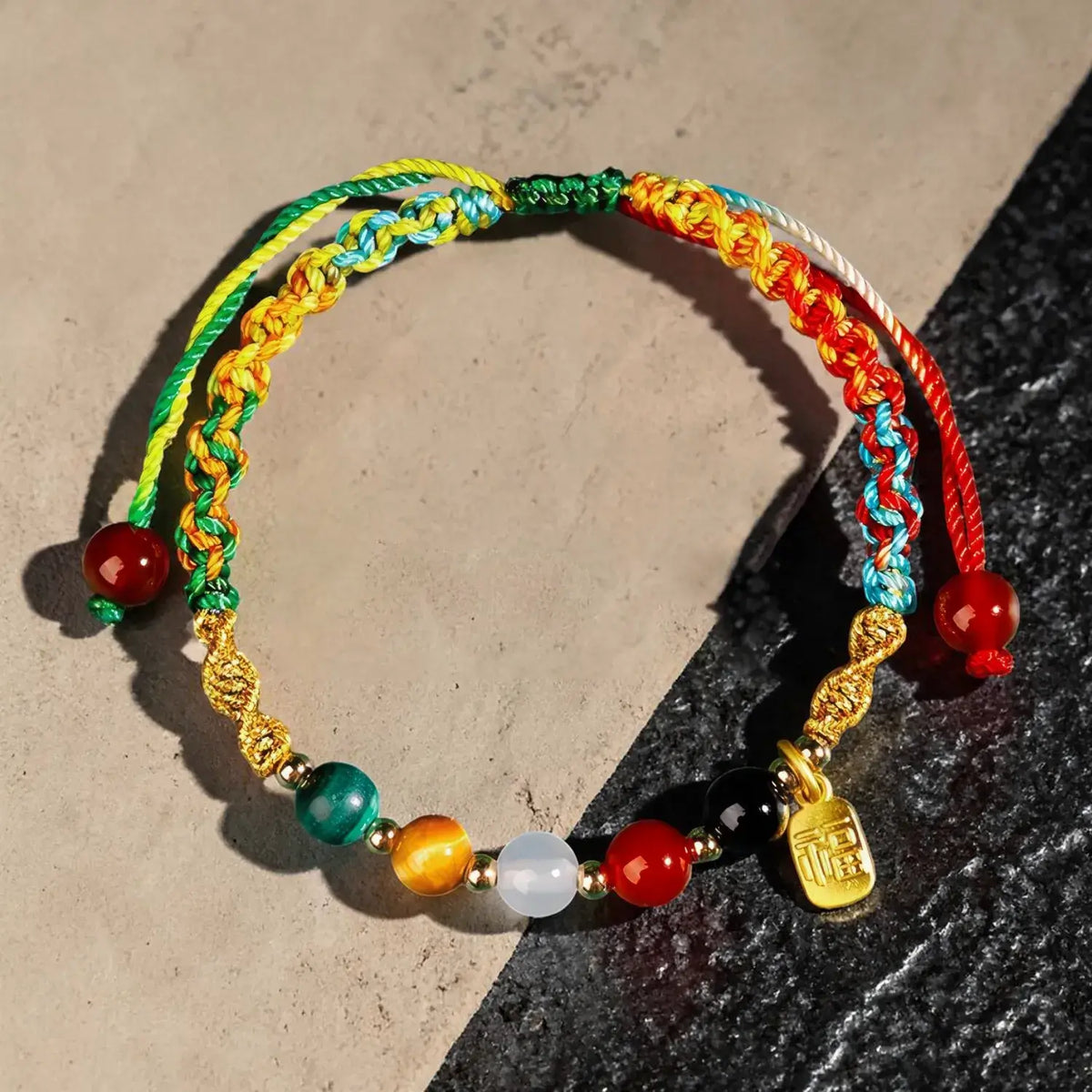 A multicolor braided cord bracelet with colorful beads and a small gold charm, displayed on a split beige and dark gray surface.