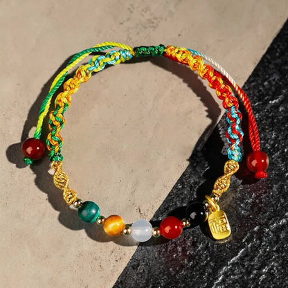 A multicolor braided cord bracelet with colorful beads and a small gold charm, displayed on a split beige and dark gray surface.