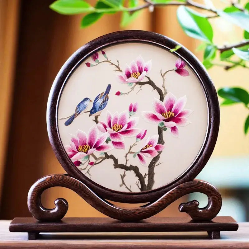 Decorative plate with floral and bird design on a wooden stand, surrounded by green leaves.
