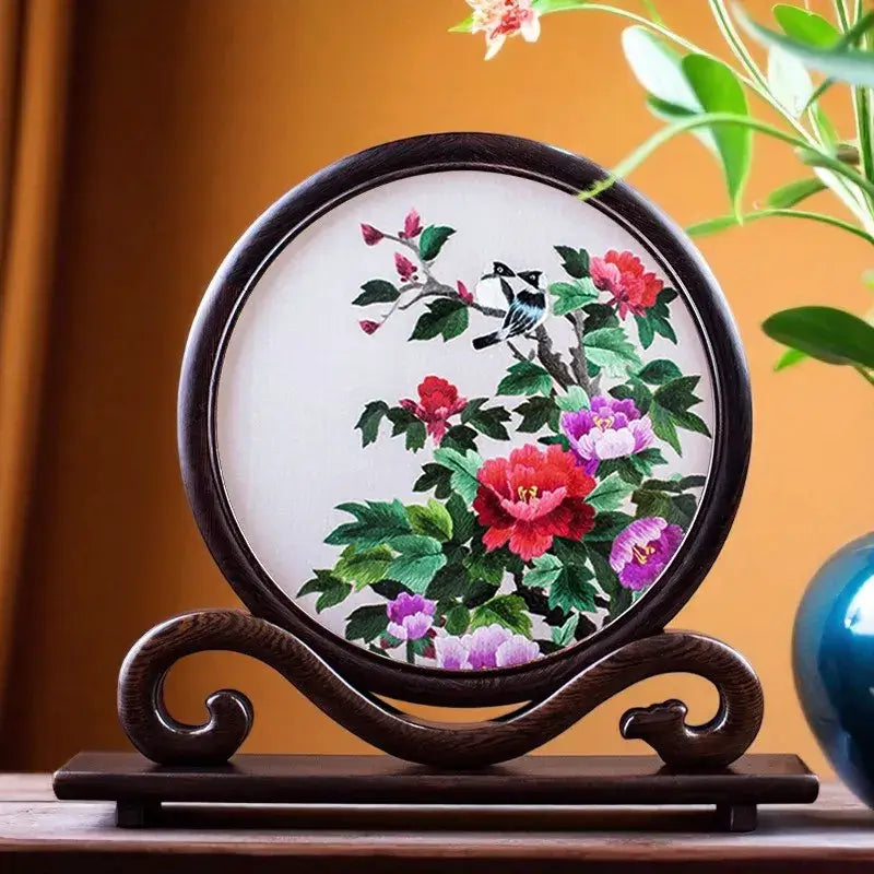Decorative embroidery of flowers and birds on a wooden stand with a plant in the background.