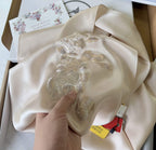 Beige fabric with gold embroidery held by a hand, with a box and card in the background.