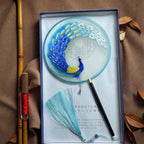 Decorative fan with peacock design in a box on a textured surface