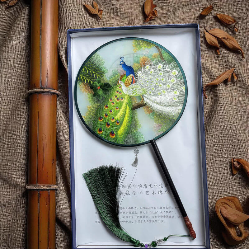 Round fan with peacock design in a box on a textured fabric background