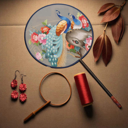 Decorative fan with peacock design, earrings, magnifying glass, spool of thread, and paintbrush on a textured surface.