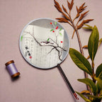 Round fan with artistic design of a bird on a branch, surrounded by leaves and a spool of thread on a beige background.