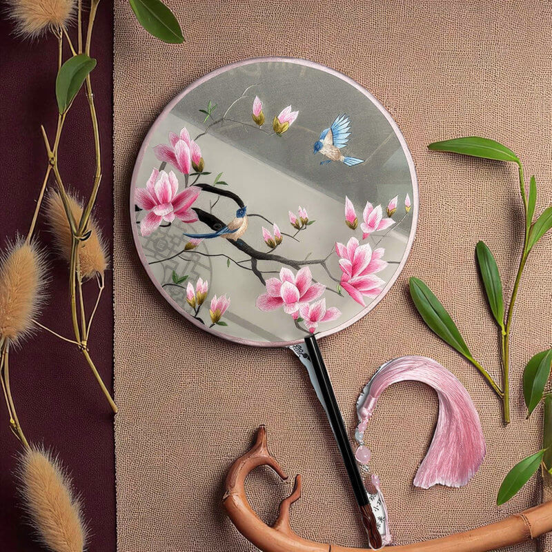 Round fan with floral and bird design on a textured surface with leaves and dried plants.