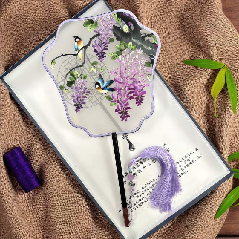 Handheld fan with purple floral design and birds, displayed on a textured surface.