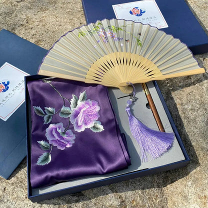 Purple embroidered fabric with floral design, wooden fan, and tassel in a box on a stone surface.