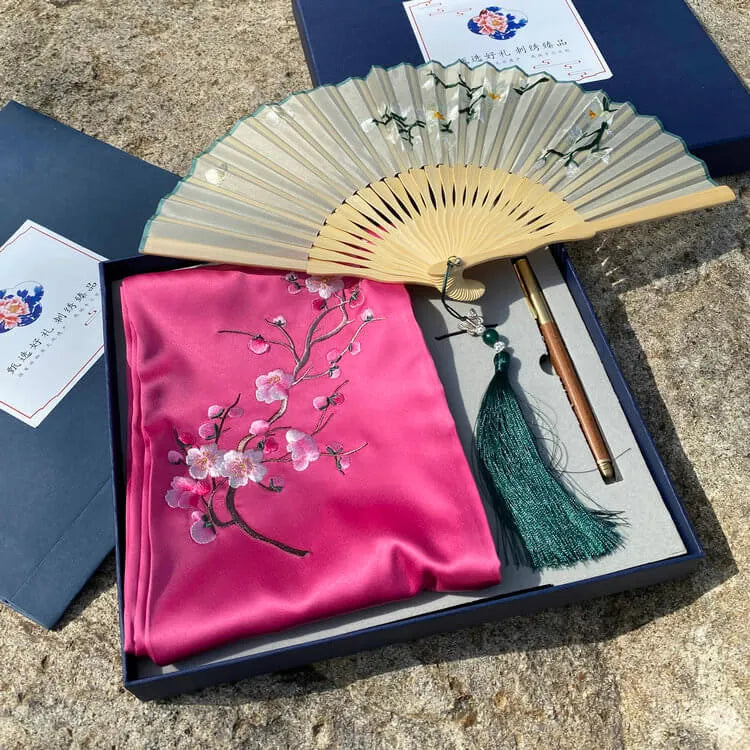 Embroidered pink fabric with cherry blossoms, a wooden fan, and a green tassel on a textured surface.